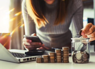 Sporiteľný účet vs. investovanie: kedy sa oplatí držať hotovosť a kedy už len míňate peniaze? Person using a smartphone beside a laptop, with stacks of coins and a coin jar on the desk indicating saving or budgeting.