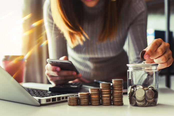 Person using a smartphone beside a laptop, with stacks of coins and a coin jar on the desk indicating saving or budgeting.