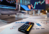 Trading vs. investovanie: Hlavné rozdiely medzi aktívnym obchodovaním a pasívnym držaním aktív Desk workspace showing stock charts on two monitors, a calculator on printed financial reports, and a smartphone in the foreground.