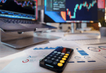 Trading vs. investovanie: Hlavné rozdiely medzi aktívnym obchodovaním a pasívnym držaním aktív Desk workspace showing stock charts on two monitors, a calculator on printed financial reports, and a smartphone in the foreground.