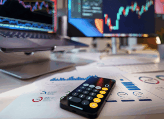 Trading vs. investovanie: Hlavné rozdiely medzi aktívnym obchodovaním a pasívnym držaním aktív Desk workspace showing stock charts on two monitors, a calculator on printed financial reports, and a smartphone in the foreground.