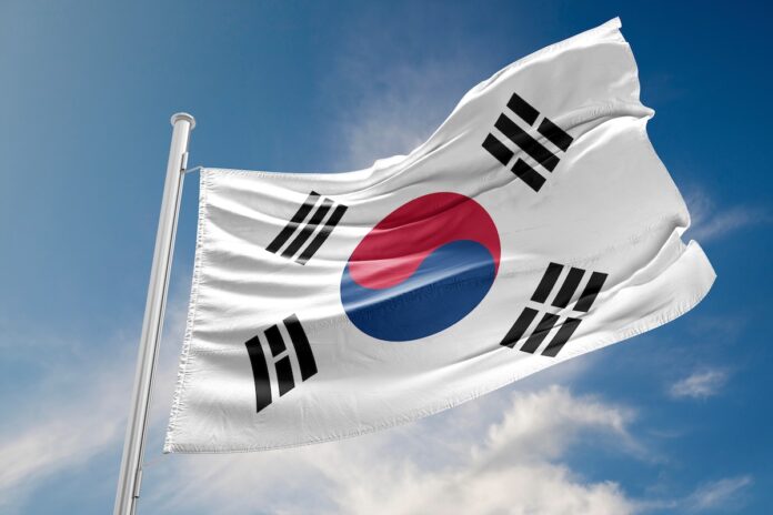 South Korean flag (Taegeukgi) flying on a flagpole against a blue sky with red-blue yin-yang and black trigrams.